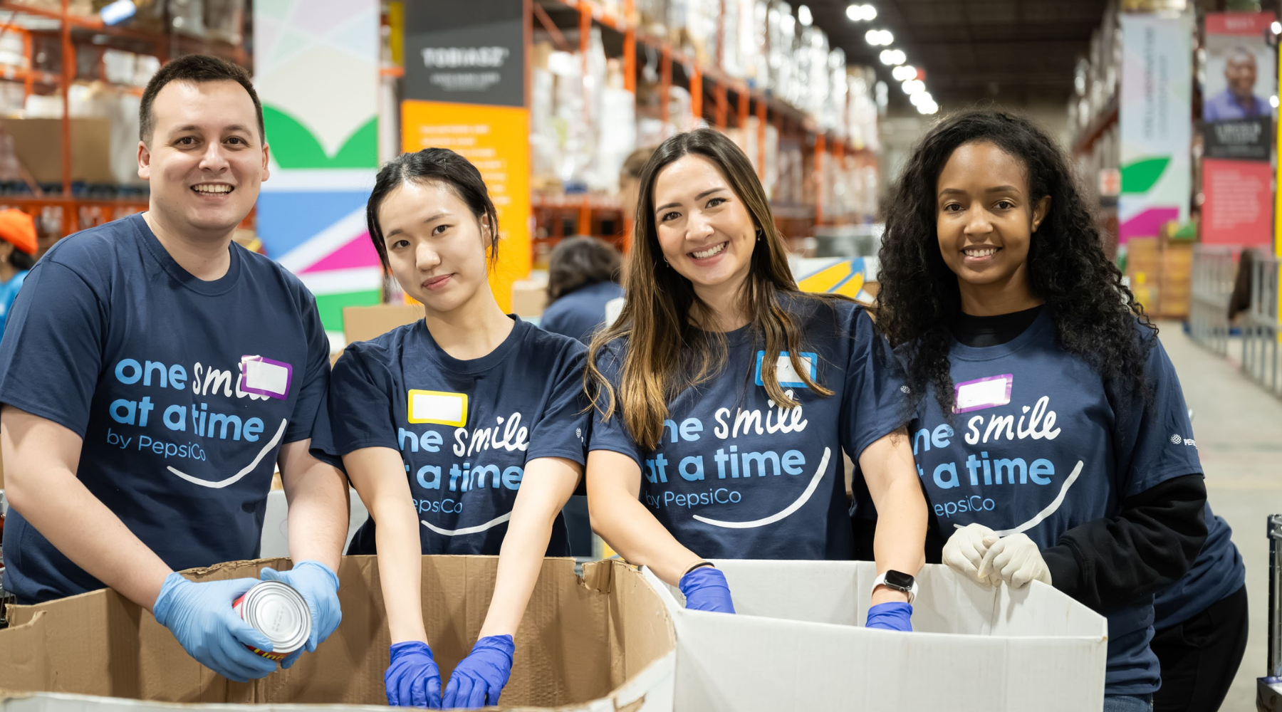 Four PepsiCo volunteers work together at a large bin in a warehouse, sorting and packing canned goods.