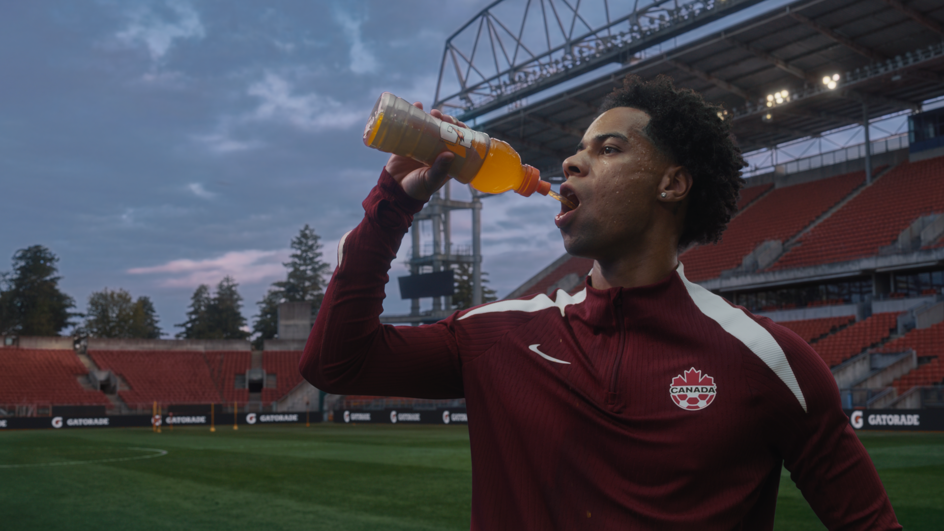 Un joueur de l'équipe canadienne de soccer vêtu aux couleurs de l'équipe nationale et buvant une boisson Gatorade orange sur le terrain d'un stade de soccer canadien, avec des panneaux de bord de terrain à l'image de Gatorade visibles en arrière-plan.